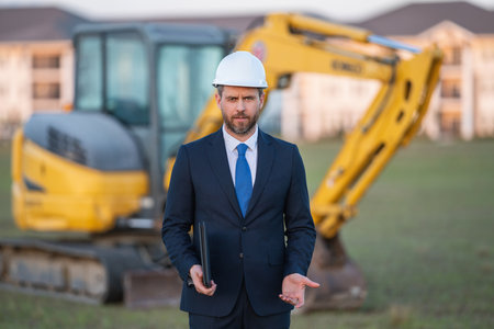 Construction manager in suit and helmet at a construction site. Construction manager worker or supervisor wearing hardhat in front of house. Supervisor construction manager near excavator. Renovation.の写真素材