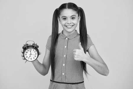 Happy teenager teen girl holding clock over yellow background. Early morning, daily schedule. Child checking time. do not be late. Punctual kid with alarm. Deadline time.の写真素材