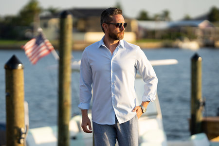 Rich summer business man near the yacht. Portrait of fashion male model outdoor. Fashion male posing near yachts. Man in urban style. Rich man at yacht club. Man in gulf with seaview and luxury ships.の写真素材