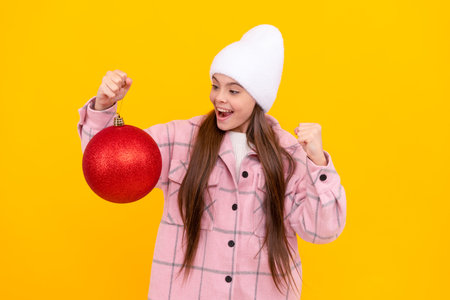 merry christmas. glad kid in winter hat. teen girl with red decorative ball on yellow background.の写真素材