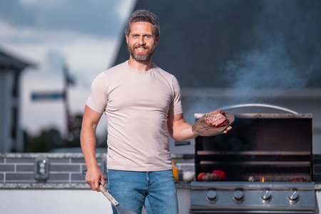 Grill cook. Chef with BBQ cooking tools. Barbecue and grill. Picnic and barbecue party. Chief cook with utensils for barbecue grill. Barbeque on holiday picnic. Man grilling a steak on BBQ.の写真素材