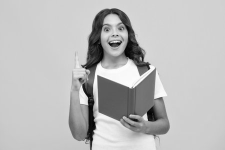 Amazed teen girl. School teenage girl with school bag backpack hold book on yellow isolated studio background. Children school and education concept. Excited expression, cheerful and glad.の写真素材