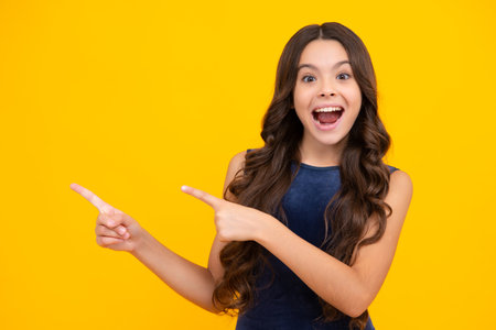 Look at advert. Teenager child points aside shows blank copy space for text promo idea presentation, poses against yellow background. Excited face, cheerful emotions of teenager girl.の写真素材