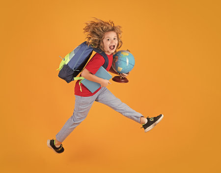 Shool kid jump with school bag and globe. Full length body of little school kid jumping having fun isolated yellow color background. Crazy school boy jump.の写真素材