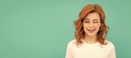winking redhead woman with curly hair on blue background. Woman isolated face portrait, banner with copy space.の写真素材