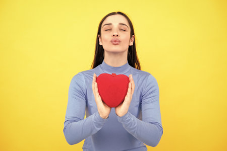 Happy woman hold red heart. Beautiful female model posing on isolated yellow background. Cheerful positive cute pretty tender girl with heart.の写真素材