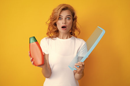 Funny girl combing hair. Redhead woman with a comb, isolated on yellow background. Woman hold bottle shampoo and conditioner. Happy young woman with balm bottle applying hair mask. Beauty product.の写真素材
