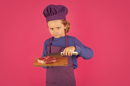 Child cook hold cutting board with meat beef steak and knife. Chef child preparing healthy food. Studio portrait of child with chef hats.の写真素材