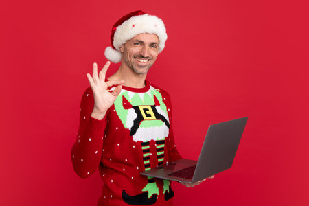 Man in holiday sweater and Santa hat hold laptop on studio background. Christmas sale or special winter offer.の写真素材