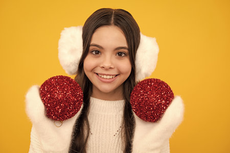 Merry christmas and happy New year. Portrait of happy teenager child wears sweater and knitted gloves on yellow isolated background. Kid in winter clothes. Teen girl with decorative christmas ball.の写真素材