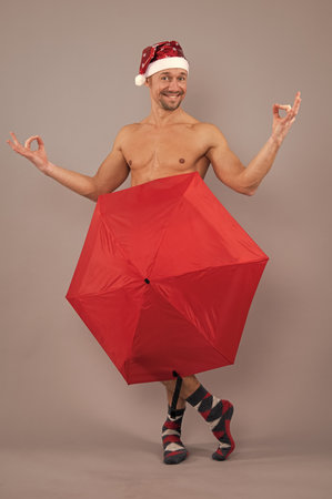 Crazy nude santa covered underwear with umbrella. Naked gay santa in santa head funny posing on gray studio background.の写真素材
