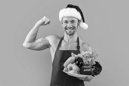 Man in cook apron and Santa hat with vegetables, healthy diet concept. Santa vegan. Young man with vegetable on gray studio background. Healthy vegan food for Christmas or new year.の写真素材