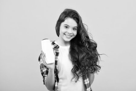 Teen child girl with shampoo bottle or shower gel isolated on yellow background. Kids hair cosmetic product. Happy teenager, positive and smiling emotions of teen girl.の写真素材