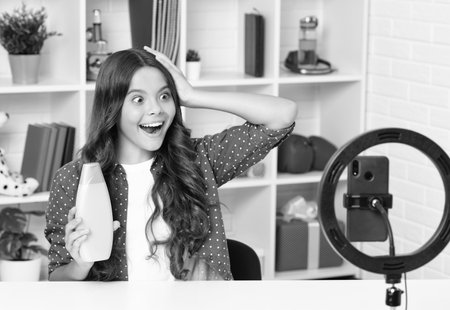 Young blogger hold hair conditioner or shampoo bottle recording video for blog. Teen girl speaking in front of camera for vlog. Teenager working as blogger, recording video tutorial for blog.の写真素材