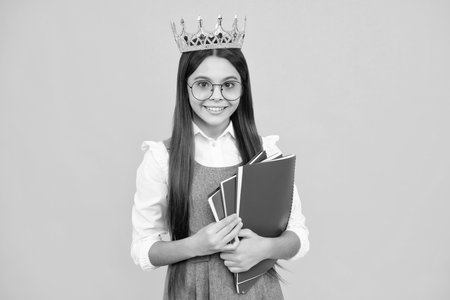 Schoolgirl in school uniform and crown celebrating victory on yellow background. School child hold books. Education graduation, victory and success. Happy teenager school girl.の写真素材