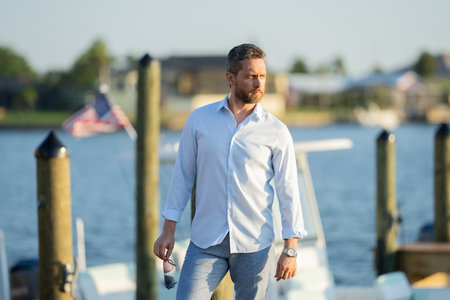 Rich summer business man near the yacht. Portrait of fashion male model outdoor. Fashion male posing near yachts. Man in urban style. Rich man at yacht club. Man in gulf with seaview and luxury ships.の写真素材