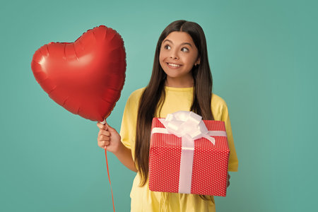 Teenager child with gift box. Present for holidays. Happy birthday, Valentines day. Kid hold present box, love valentines heart balloon. Happy girl face, positive and smiling emotions.の写真素材