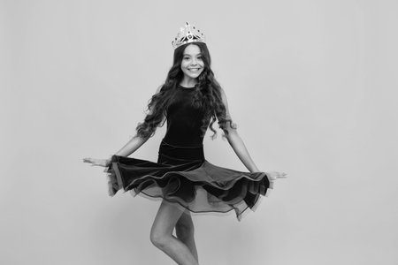 Movement windy dress. Teenage selfish girl celebrates success victory. Teen child in queen crown isolated on yellow background. Princess in tiara. Prom party.の写真素材