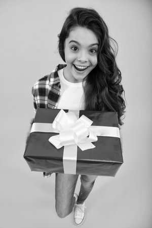 Teenager child in shirt holding gift box on yellow isolated background. Gift for kids birthday. Happy teenager, positive and smiling emotions of teen girl.の写真素材