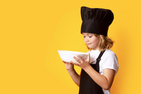 Funny child cook with cooking plate. Excited chef cook. Child wearing cooker uniform and chef hat preparing food, studio portrait.の写真素材