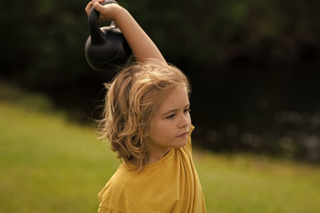 Kid lifting the kettlebell in park outside. Child boy raising a kettlebell. Cute child training with dumbbells. Kids fitness. Kid boy exercising with dumbbells. Healthy kids.の写真素材