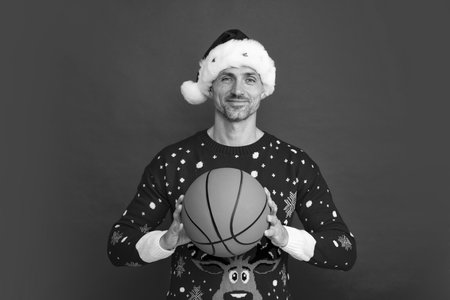Man trainer hold basketball ball in holiday sweater and Santa hat on studio background. Christmas sale or special winter offer.の写真素材