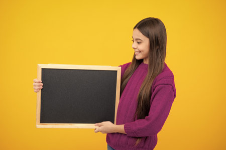 Teenage girl child holding blackboard, isolated on a yellow background.