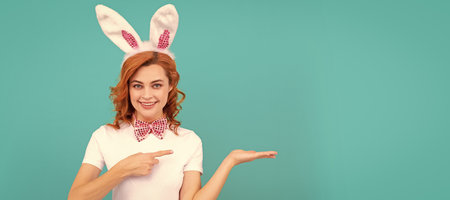 Easter bunny girl. Woman isolated face portrait, banner with mock up copyspace. happy easter woman in bunny ears and bow tie presenting product and point finger on blue background.の写真素材