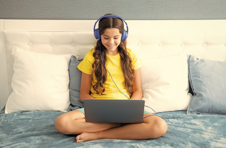 Happy teenage child in headphones relax on bed at home listen to music using laptop. Child in earphones browse internet on computer, friendship communication.の写真素材