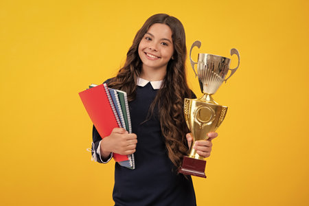 Teenager school girl with award winner trophy. Child hold books with gold trophy or winning cup isolated on yellow. Education graduation, victory and winning.の写真素材