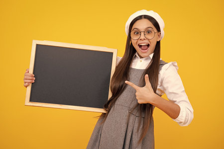 Teenager younf school girl holding school empty blackboard isolated on yellow background. Portrait of a teen female student. Excited teenager girl.の写真素材