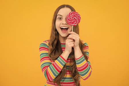 Cool teen child with lollipop over yellow isolated background. Sweet childhood life. Teen girl with yummy lollipop candy. Excited face, cheerful emotions of teenager girl.の写真素材