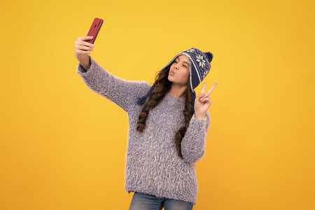 Kids selfie. Close-up portrait of cute teen girl in winter knitted hat using mobile phone, isolated over bright vivid vibrant yellow color background. Funny face.の写真素材