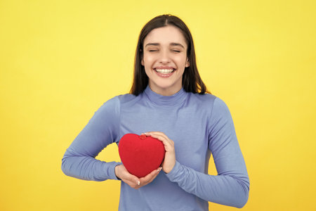Beautiful girl holding valentines gift on yellow background. Portrait of young woman holding red paper heart.の写真素材