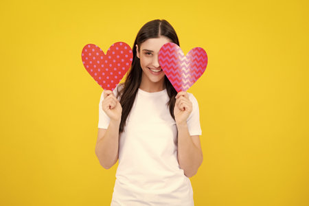 Young woman valentines day concept, girl with heart shaped card. Lovely charming girl holding in hands heart love symbol isolated on yellow background.の写真素材