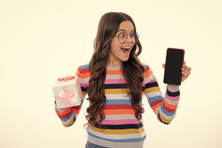 Birthday present, bonus for mobile user. Portrait of teenager child girl holding gift box and cell phone with mockup cipy space blank display screen. Studio shot isolated on white background.の写真素材