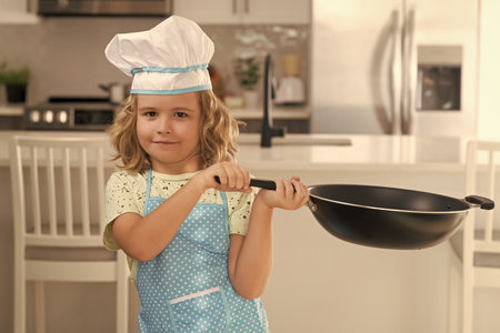 Kid chef cook cookery with pan in kitchen. Fun kids kitchen. Funny little kid chef cook wearing uniform cook cap and apron cooked food in the kitchen. Kids are preparing food, bake cookies.の写真素材