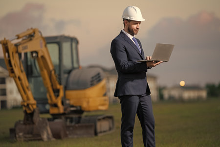 Architect at a construction site. Architect man in helmet and suit at modern home building construction. Architect with a safety vest and suit. Confident architect standing at house background.の写真素材