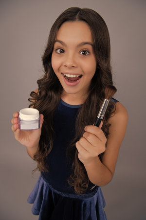 Beauty, make up and kids cosmetics. Teenage girl 12, 13, 14 years old with brush applying blush to face isolated on gray background. Excited face, cheerful emotions of teenager girl.の写真素材