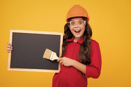 Painter in workshop. Renovation and repair. Teen girl in helmet with painting brush. Child in hard hat. Kid builder painter on construction site. Excited teenager girl.の写真素材