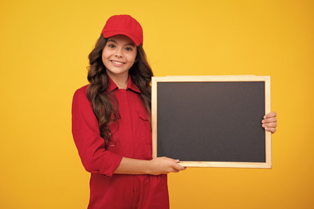 Child girl builder in red building uniform and cap. Teenage girl worker hold blackboard isolated on yellow background. Kids renovation concept. Copy space on empty board, mock up for menu.の写真素材