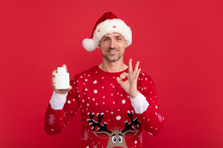 Man in Christmas sweater and Santa hat hold bottle of medicine on red background.の写真素材