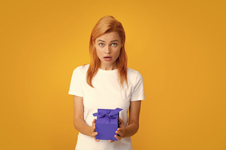 Surprised woman holding gift. Beautiful young woman in shirt hold present box with gift ribbon bow isolated on bright yellow background, studio portrait. Birthday concept.の写真素材