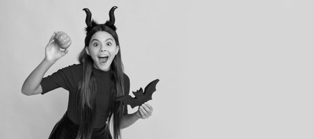 amazed kid with pumpkin and bat wearing devil horns on yellow background. Halloween kid girl portrait, horizontal poster. Banner header with copy space.の写真素材