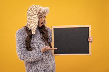 Teenage girl child in warm winter hat and sweater holding blackboard, isolated on a yellow...