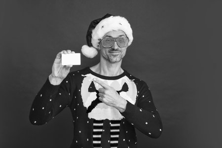 Santa man hold card. Handsome man with party glasses in a winter sweater and Santa hat on studio red background.の写真素材