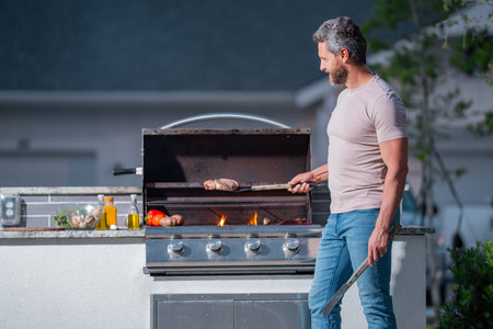 Barbecue chef. Middle aged hispanic man in apron for barbecue. Roasting and grilling food. Man hold cooking utensils barbecue. Roasting meat outdoors. Barbecue and grill. Cooking meat in backyard.の写真素材