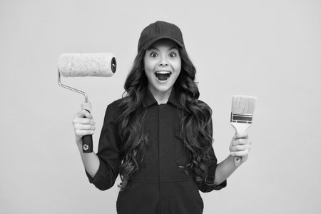 Excited teenager girl builder in building uniform and cap. Painter with painting brush tool or paint roller. Worker isolated on yellow background. Kids renovation concept.の写真素材