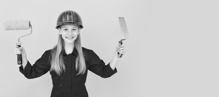 Architect girl. smiling teen child in builder hard hat with paint roller and spatula on yellow wall. Child builder in helmet horizontal poster design. Banner header, copy space.の写真素材