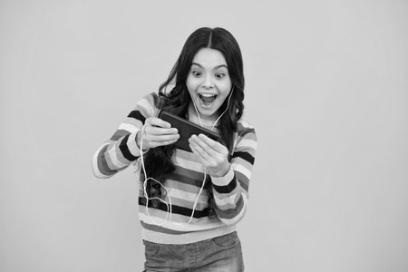 Amazed teenager use smart phone, share social media, chatting by mobile phone wear stylish casual trendy clothes isolated over yellow background. Child with smartphone. Excited teen girl.の写真素材